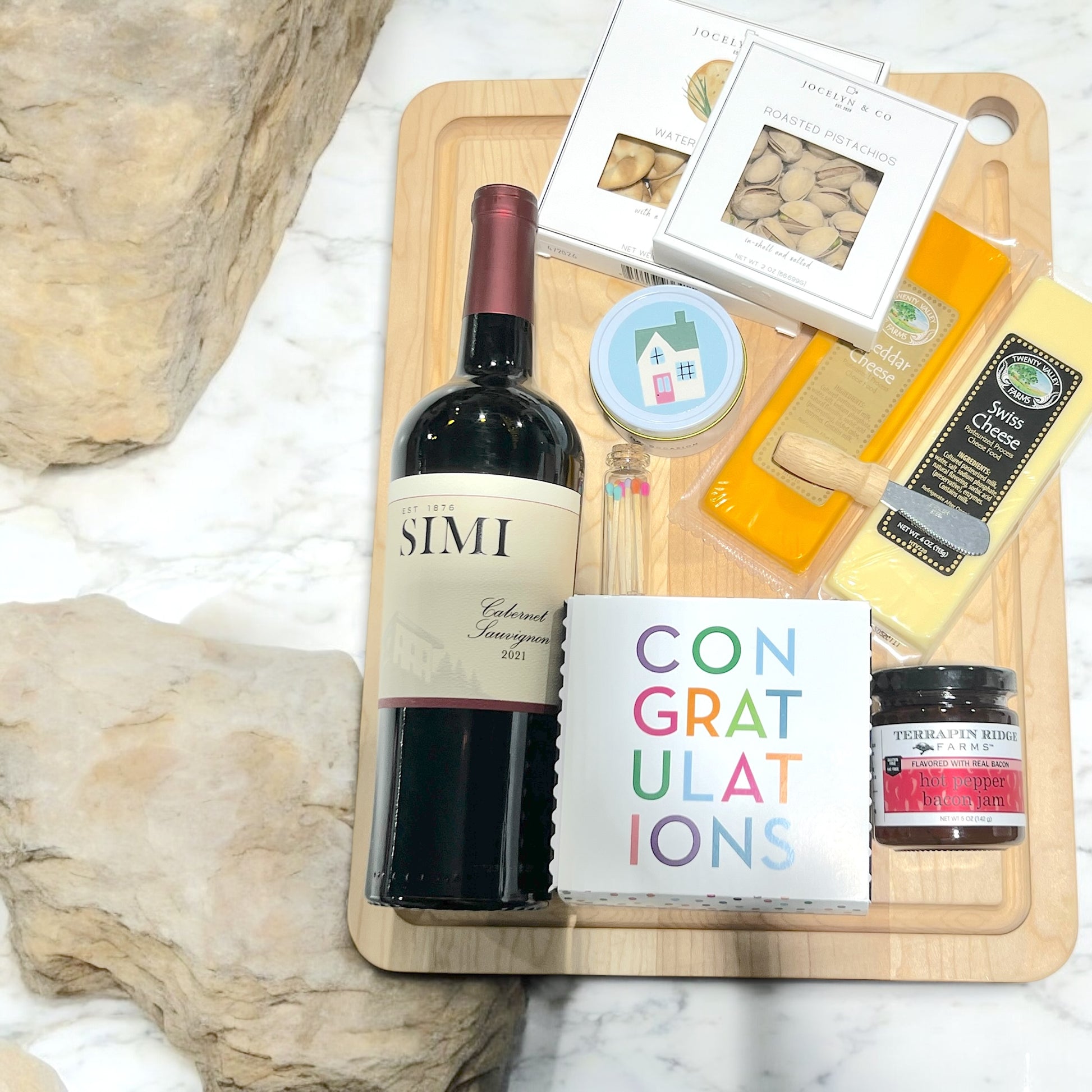 A beautifully arranged cutting board featuring a bottle of wine alongside assorted cheeses, a cutting knife, a house candle, congratulatory cookies, crackers, nuts, and jam. This gourmet gift set ships nationwide from New York City.