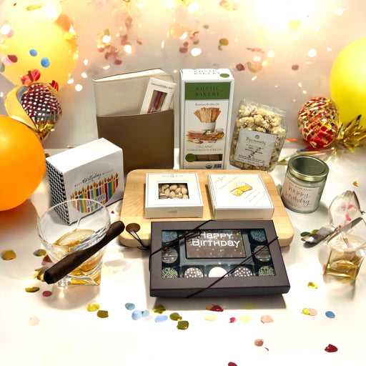 A birthday gift basket filled with delicious treats including cookies, chocolates, crackers, cheese, popcorn; accompanied by a journal, a candle, and a whiskey glass with a cigar holder.