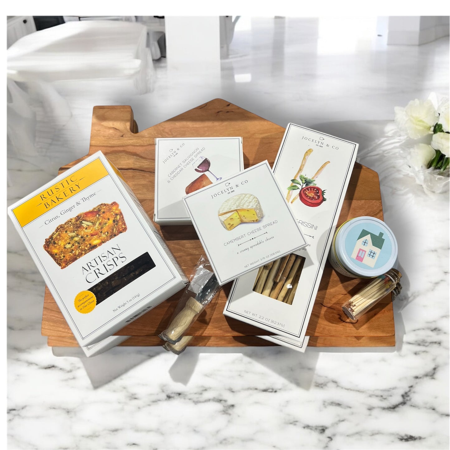 Cozy cabin cutting board with assorted crackers and cheeses, house candle and cutting knife. Makes a great housewarming gift.