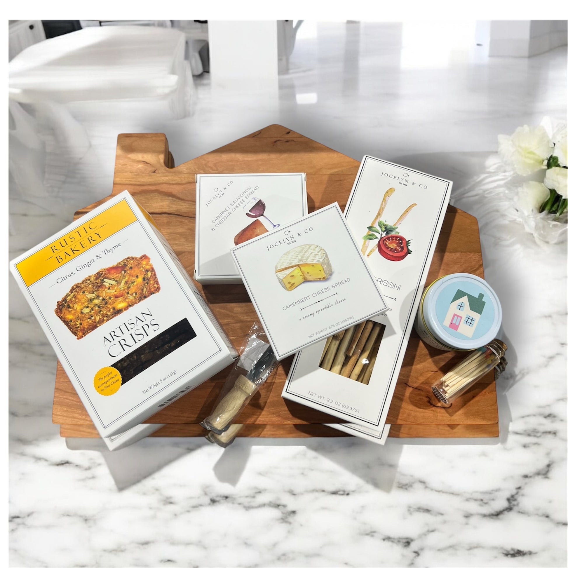 Cozy cabin cutting board with assorted crackers and cheeses, house candle and cutting knife. Makes a great housewarming gift.
