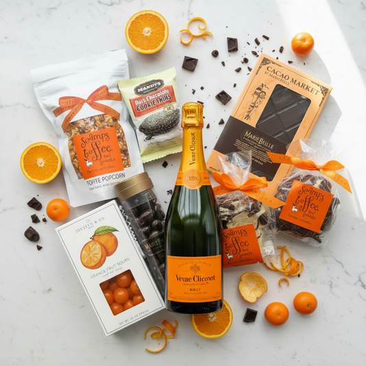 A luxury gift basket featuring an orange and citrus-themed champagne bottle, assorted chocolates, and packaged snacks from Scavengers of the Fleece and Cacao Market, styled on a white marble background with fresh orange slices and peels.