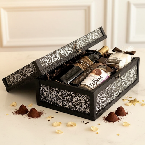 A black wooden gift box with ornate white damask patterns, shown open to reveal an assortment of chocolates and treats inside. The box sits on a light cream marble surface with scattered chocolate pieces and gold star confetti around it.