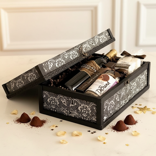 A black wooden gift box with ornate white damask patterns, shown open to reveal an assortment of chocolates and treats inside. The box sits on a light cream marble surface with scattered chocolate pieces and gold star confetti around it.