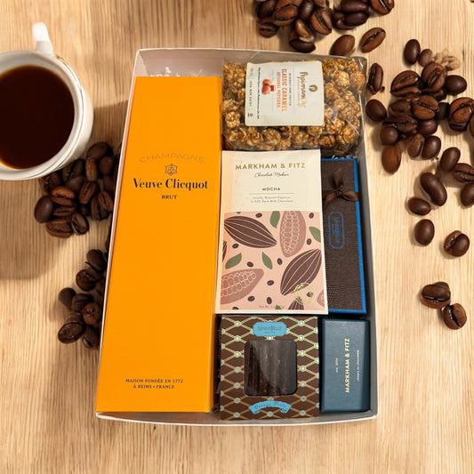 Box of chocolate bars with a Veuve Cliquot bottle on a wooden surface with coffee beans and a cup of coffee.