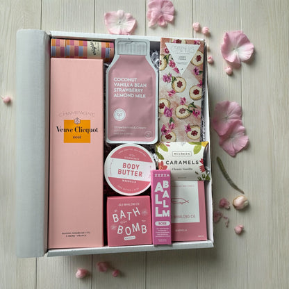 Gift box with various items including a bottle of Veuve Clicquot, a bath bomb, and a body butter on a wooden surface with pink flowers.