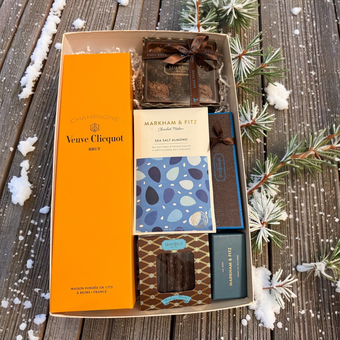 Gift box with Veuve Clicquot bottle, Markham & Fitz chocolate, and other items on a wooden surface with snow and pine branches.
